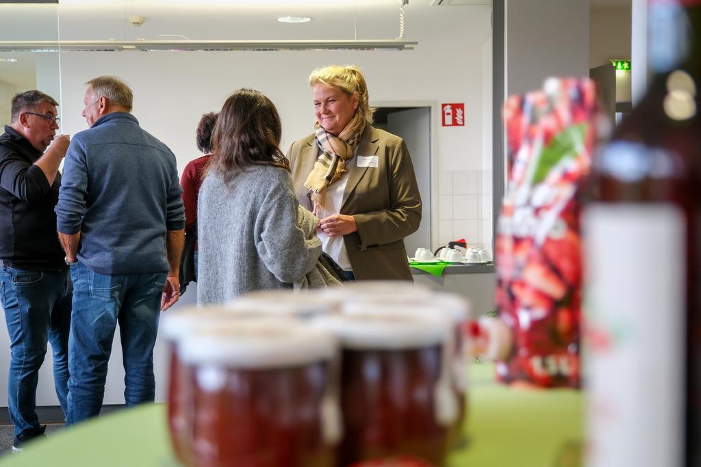 Menschen stehen in einem Raum und unterhalten sich, im Vordergrund stehen Gläser mit roter Marmelade und eine Flasche.
