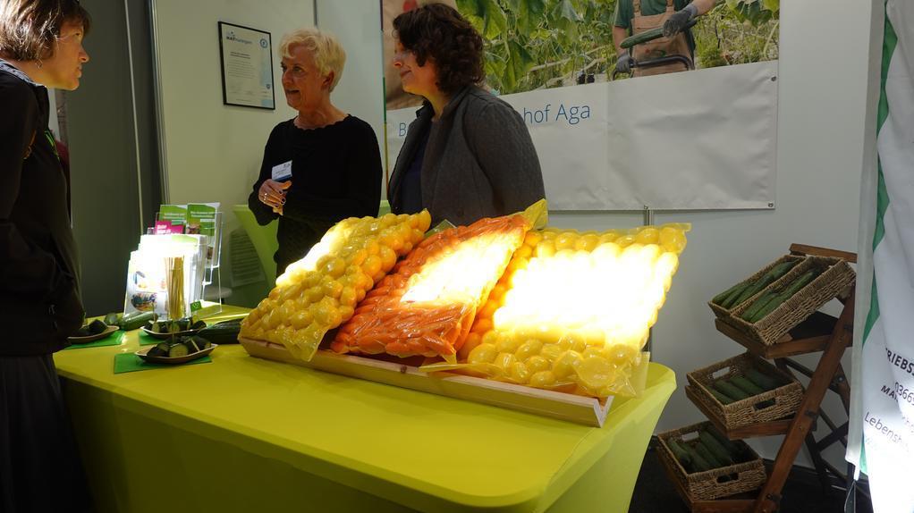 Drei Personen stehen an einem gelb gedeckten Tisch mit drei Tabletts voller gelber und oranger Lebensmittel in einem Innenraum mit Pflanzenposter im Hintergrund