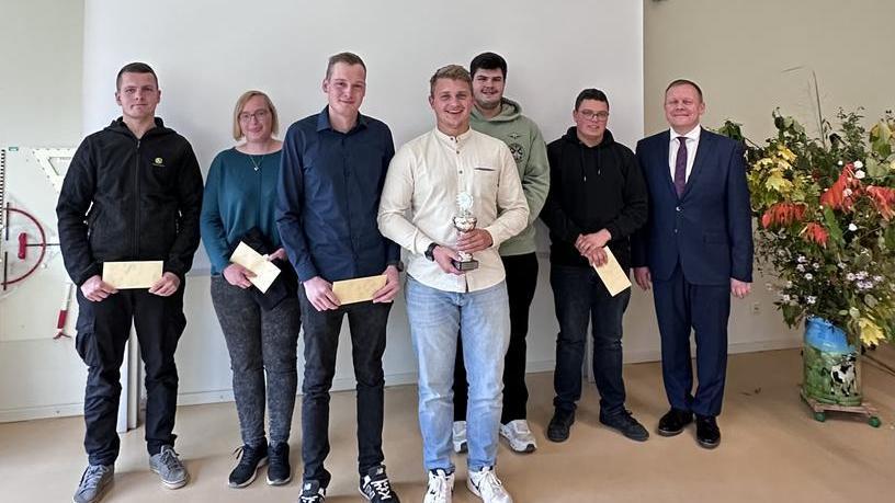 Sieben Personen stehen nebeneinander in einem Raum, eine hält einen Pokal, andere halten Umschläge, rechts steht ein Mann im Anzug, links eine große Blumendekoration.