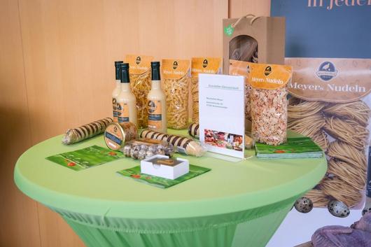 Rund gedeckter Tisch mit grüner Tischdecke, darauf verschiedene Nudelsorten, Flaschen mit Soße, Verpackungen und ein Informationsblatt.