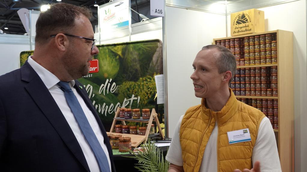 Zwei Männer mit Namensschildern stehen an einem Messestand mit Produktproben und Preistafeln für Fruchtaufstriche und Kurds.
