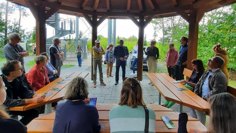 Eine Gruppe von Menschen versammelt sich unter einem Holzpavillon. Einige stehen, andere sitzen an langen Holztischen. Im Hintergrund sind Bäume und eine Treppe sichtbar.