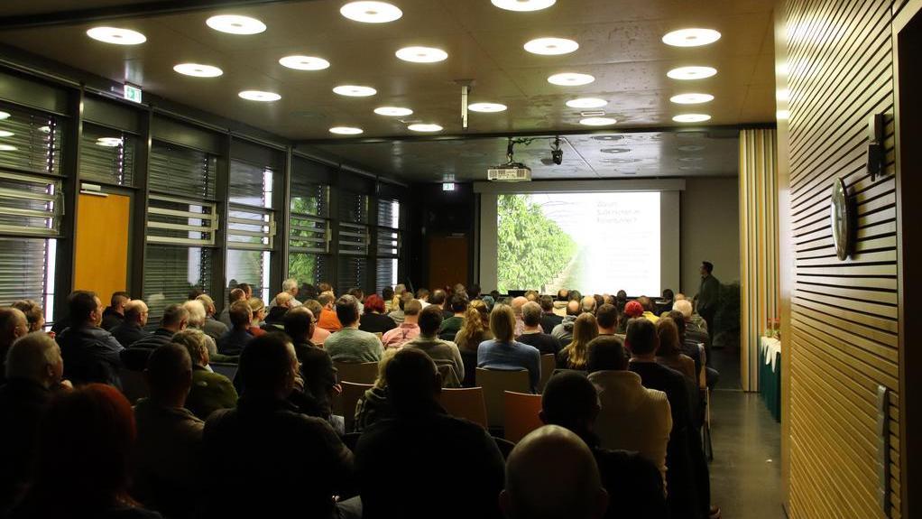 Publikum sitzt in einem dunklen Raum vor einer großen Leinwand mit einer Präsentation
