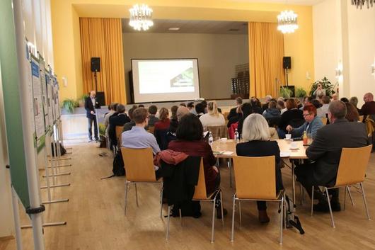 Person hält Vortrag vor Publikum in Saal mit Holzfußboden, gelben Vorhängen und Kronleuchtern, Zuschauer sitzen an Tischen.