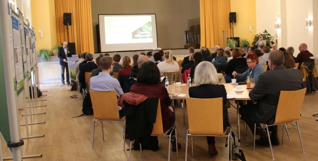 Person hält Vortrag vor Publikum in Saal mit Holzfußboden, gelben Vorhängen und Kronleuchtern, Zuschauer sitzen an Tischen.