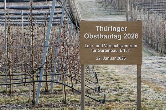 Holzschild mit der Aufschrift 'Thüringer Obstbautag 2026 Lehr- und Versuchszentrum für Gartenbau, Erfurt 22. Januar 2026' vor kahlen Obstbäumen und Netzen in einem frostigen Obstgarten