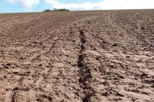 Braunfarbener, trockener Ackerboden mit einer tiefen, schmalen Erosionsrinne in der Mitte unter blauem Himmel.