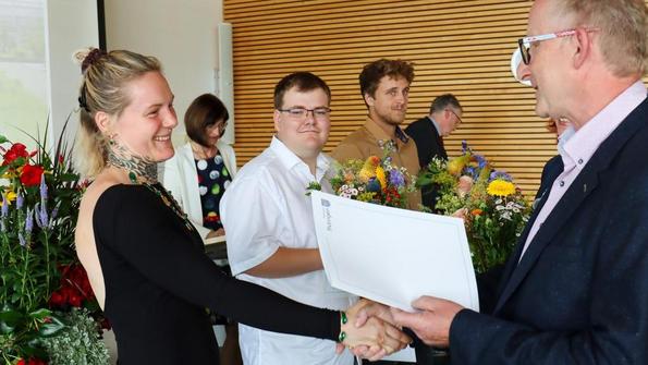 Übergabe eines Zeugnisses an eine Absolventin bei einer feierlichen Veranstaltung mit Blumenarrangements.