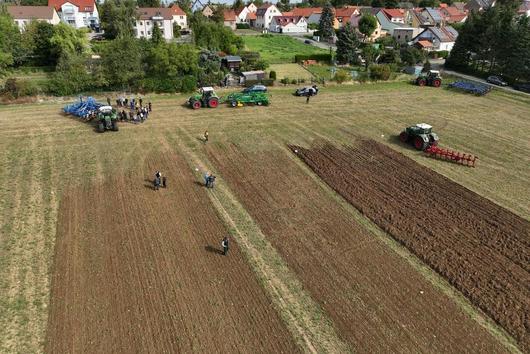 Das Bild zeigt eine Luftaufnahme eines Feldes, auf dem verschiedene landwirtschaftliche Maschinen und Geräte ausgestellt sind. Mehrere Gruppen von Personen stehen in der Nähe der Maschinen und scheinen sich die Geräte anzusehen oder eine Demonstration zu beobachten. Im Hintergrund ist ein Wohngebiet zu erkennen. Das Feld selbst zeigt unterschiedlich bearbeitete Bereiche, möglicherweise um verschiedene Bodenbearbeitungsmethoden zu vergleichen. Es wirkt wie eine Art Feldversuch oder eine Vorführung landwirtschaftlicher Technik.