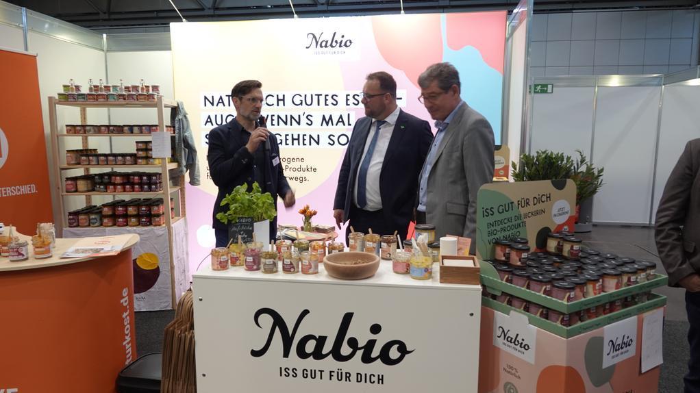 Drei Männer in Anzügen stehen an einem Messestand mit dem Schriftzug 'Nabio ISS GUT FÜR DICH' und verschiedenen Gläsern und Produkten auf dem Tisch