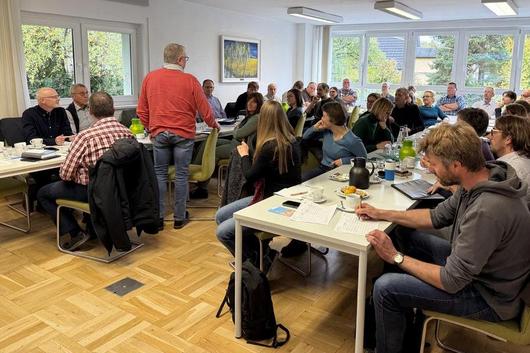 Mehrere Personen sitzen an Tischen in einem hellen Raum mit großen Fenstern, einige schreiben, andere hören zu.