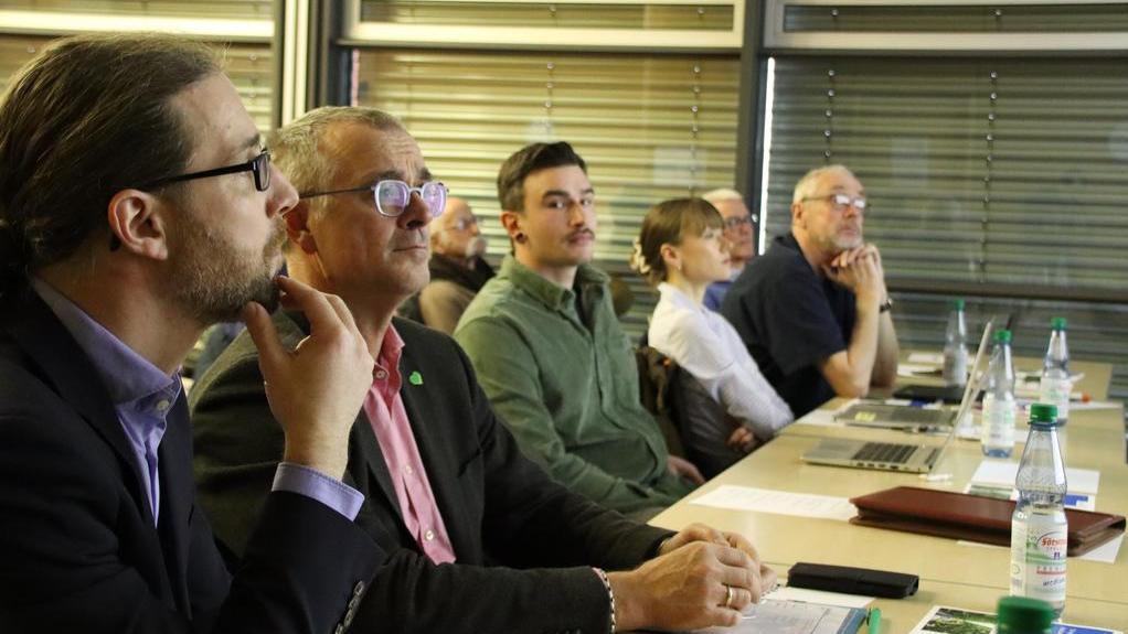 Mehrere Personen sitzen in einer Reihe an einem Tisch mit Laptops und Wasserflaschen und blicken nach vorne