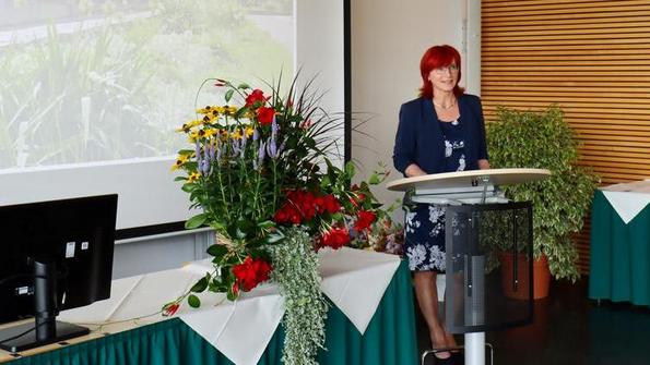 Frau in blauem Blazer und geblümtem Kleid steht am Rednerpult in einem Raum mit Holzvertäfelung, Blumenarrangements und Projektionsleinwand mit Text 'Zeugnisübergabe Fachschule Gartenbau 18.07.2025'.