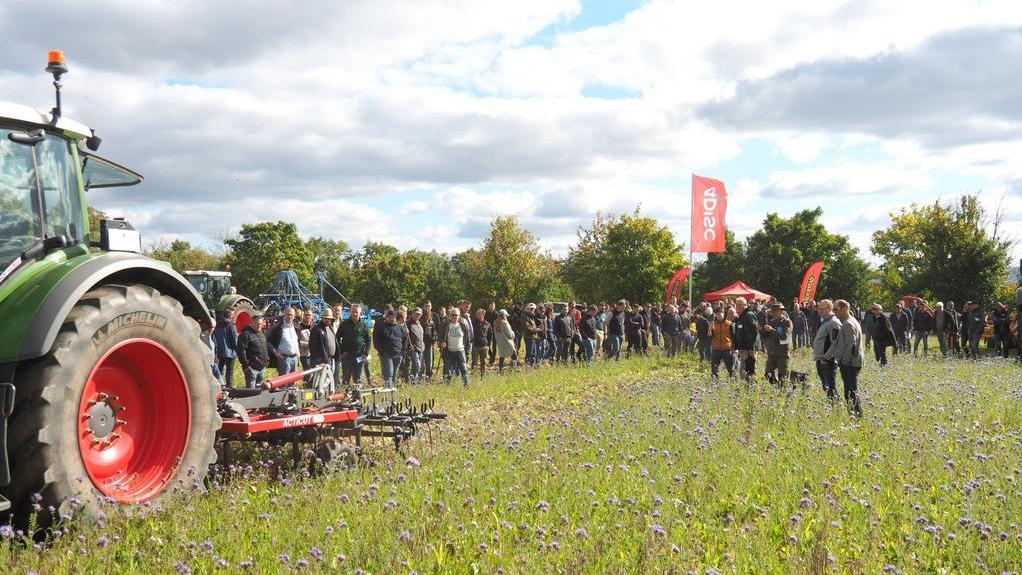 Große Menschenmenge steht auf einer Wiese mit lila Blüten, links ein Traktor mit rotem Reifen, im Hintergrund rote Fahnen.