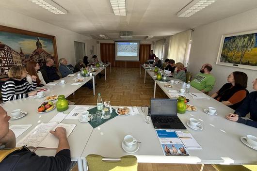 Menschen sitzen an zwei langen Tischen in U-Form in einem hellen Raum und hören einer Person zu, die vor einer Leinwand mit Präsentation steht