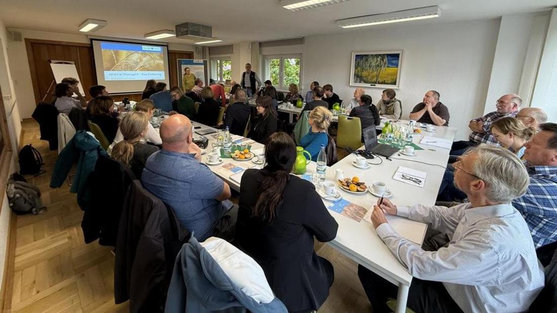 Menschen sitzen an zwei langen Tischen in einem hellen Raum, vorne steht eine Person vor einer Leinwand mit Präsentation.