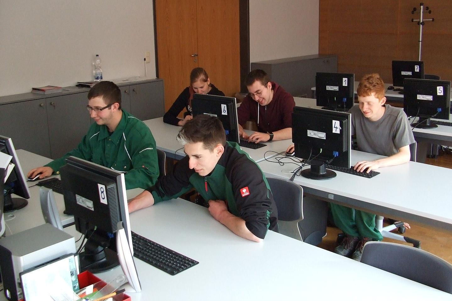 Mehrere Personen sitzen an Tischen mit Desktop-Computern in einem Klassenraum mit Holzverkleidung an der Wand.
