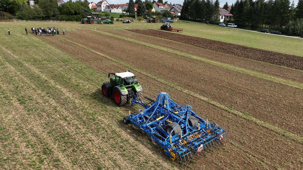 Das Bild zeigt eine Luftaufnahme eines Feldes, auf dem ein grüner Traktor mit einer blauen Saatmaschine arbeitet. Im Hintergrund sind einige Häuser und weitere landwirtschaftliche Maschinen zu sehen. Eine Gruppe von Menschen steht am Rande des Feldes und beobachtet die Arbeiten. Das Bild vermittelt den Eindruck einer Demonstration oder eines Feldtages für landwirtschaftliche Technik. Die Bearbeitung des Bodens wird dargestellt, möglicherweise zur Vorbereitung der Aussaat.