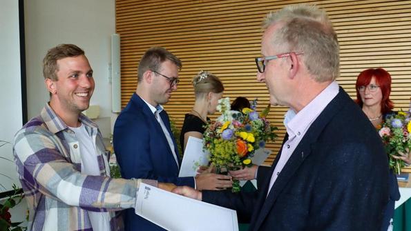 Mann mit Brille und Sakko übergibt Urkunde an jungen Mann in kariertem Hemd, im Hintergrund Personen mit Blumensträußen.