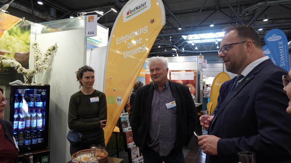 Mehrere Personen stehen an einem Messestand mit orangen Fahnen, auf denen 'Erfrischung ohne Umweg' steht, und einem Kühlschrank mit Getränkeflaschen.