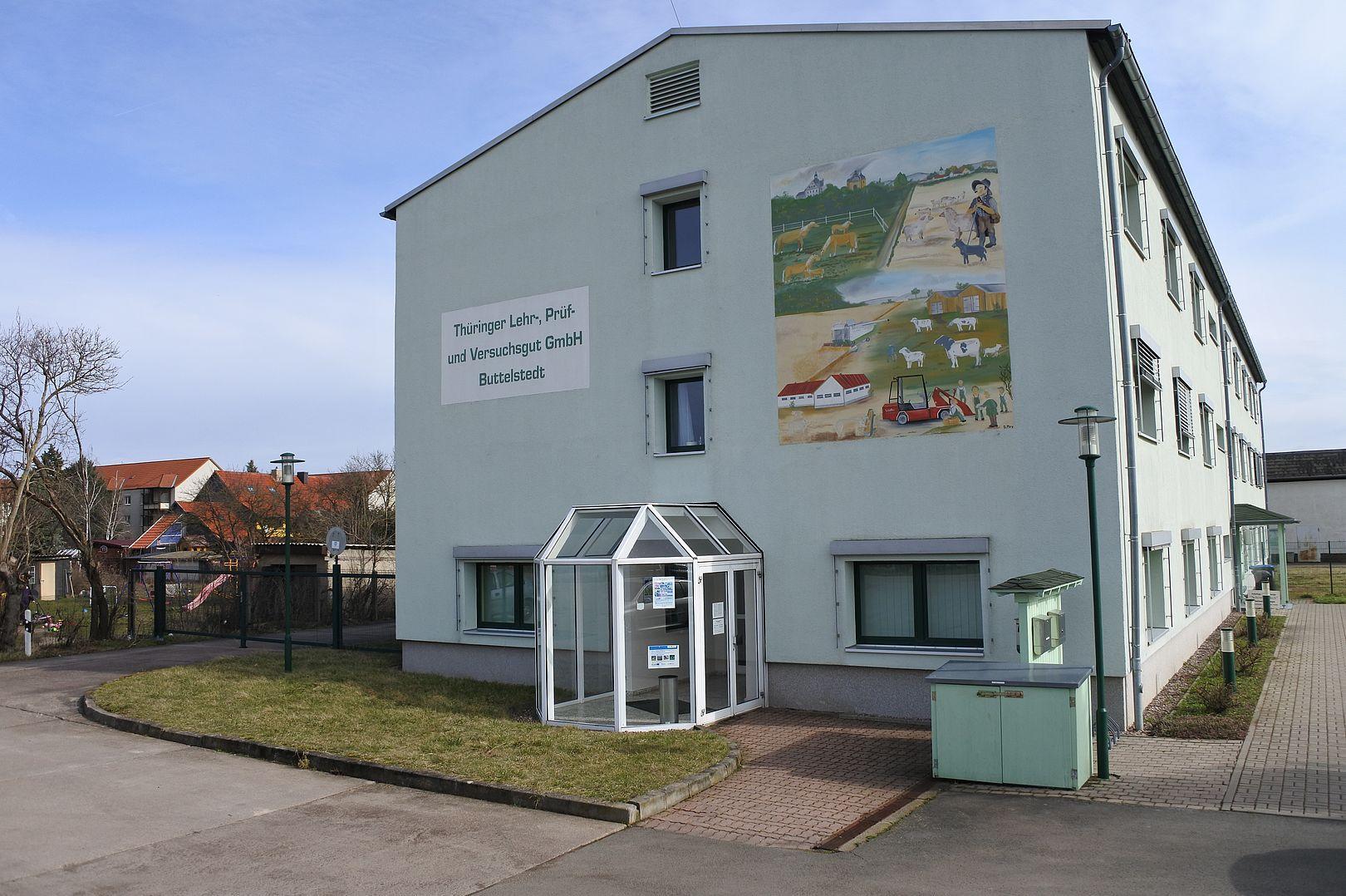 Mehrstöckiges Gebäude mit weißer Fassade, auf der ein großes Wandbild und ein Schild mit der Aufschrift 'Thüringer Lehr-, Prüf- und Versuchsstätte GmbH Buttelstedt' zu sehen sind.