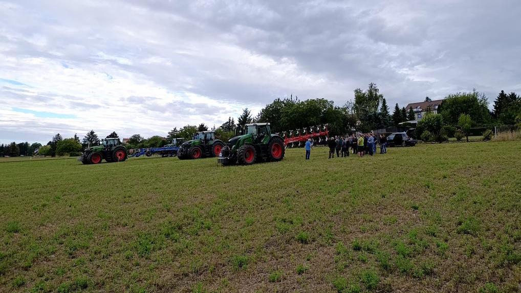 Das Bild zeigt mehrere grüne Traktoren mit angehängten Pflügen auf einem Feld. Eine Gruppe von Menschen steht vor den Traktoren und scheint sich diese anzusehen. Im Hintergrund ist etwas Bebauung und etwas Bewuchs zu erkennen. Der Himmel ist bewölkt. Es handelt sich vermutlich um eine Vorführung von Landwirtschaftstechnik.