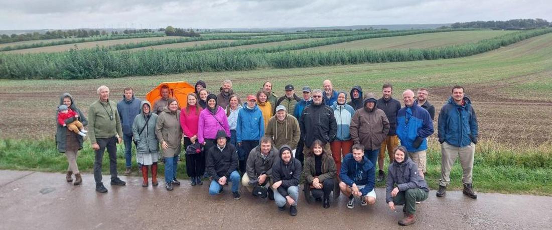 Gruppe von 28 Personen, teils stehend, teils kniend, auf einem Feldweg mit landwirtschaftlichen Flächen und Bäumen im Hintergrund, einige tragen bunte Jacken und eine Person hält einen orangefarbenen Regenschirm.