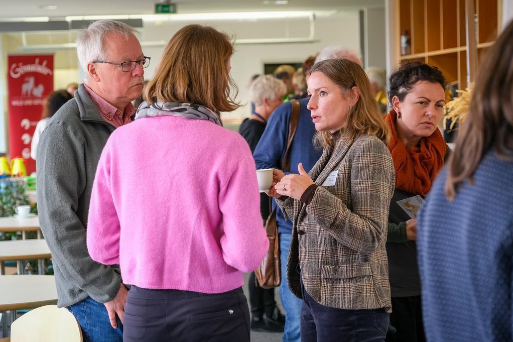 Menschen in einem Raum unterhalten sich, eine Frau trägt einen pinkfarbenen Pullover, eine andere eine karierte Jacke.