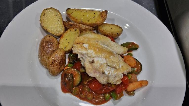 Weißer Teller mit gebratenem Fischfilet auf Gemüsemischung und gebackenen Kartoffelspalten auf Edelstahloberfläche.