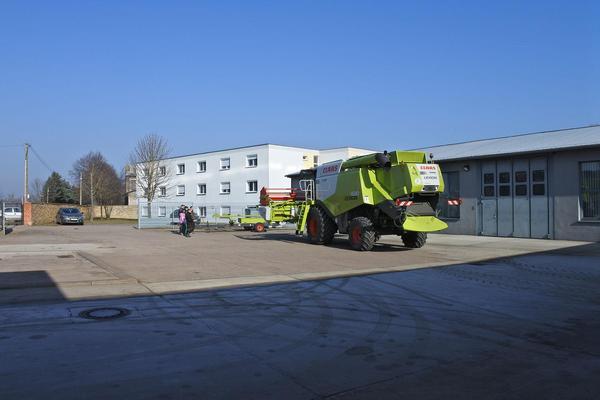 Großer grüner Mähdrescher auf Asphaltfläche vor weißem Industriegebäude mit mehreren Fenstern und Garagentoren.