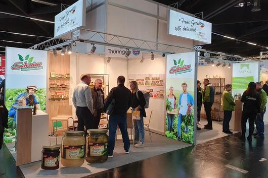 Messestand mit "Schweizer"-Markenwerbung, mehreren Personen und Produktgläsern auf dem Boden
