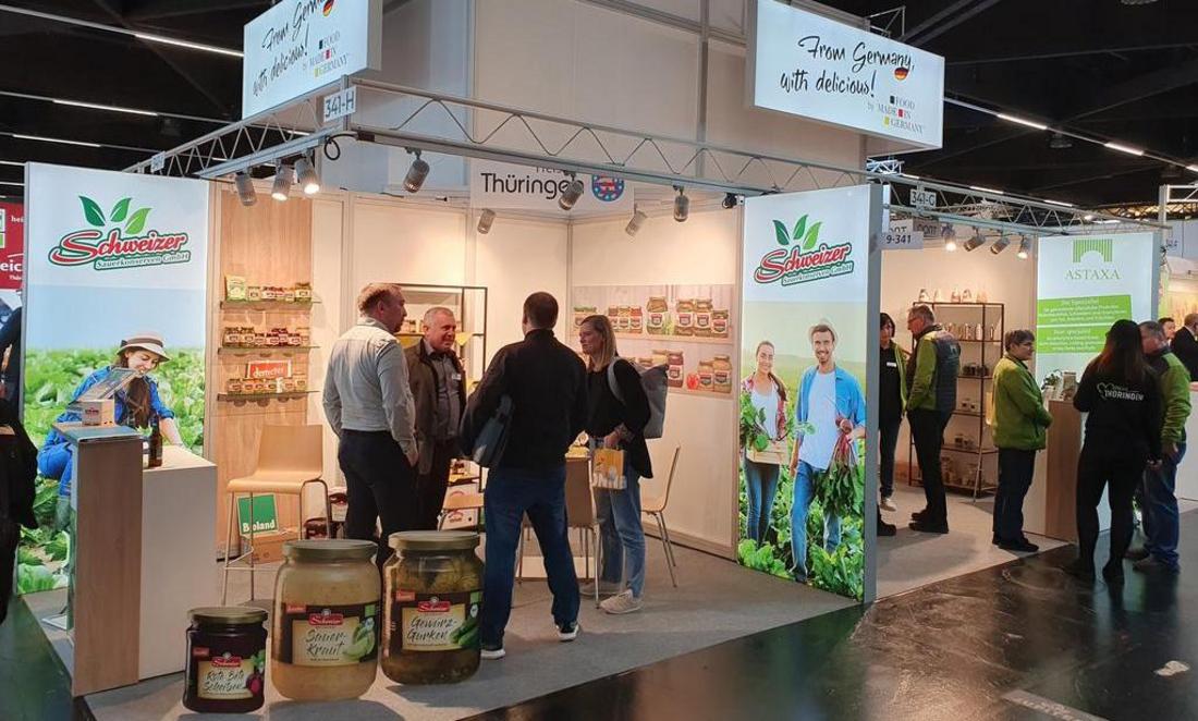 Messestand mit "Schweizer"-Markenwerbung, mehreren Personen und Produktgläsern auf dem Boden