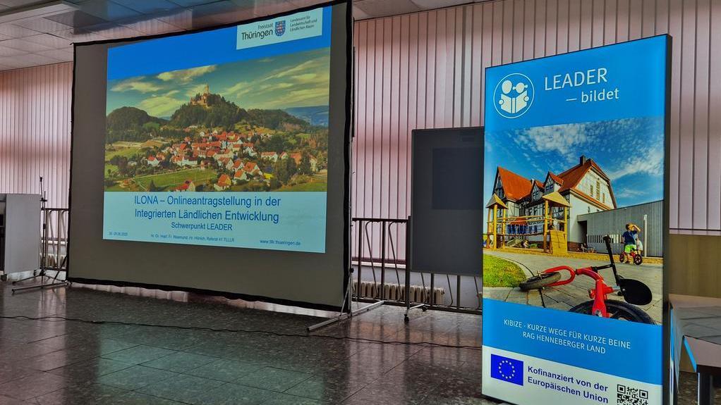 Präsentationsleinwand mit einer Landschaftsaufnahme und dem Text 'ILONA - Onlineveranstaltung in der integrierten ländlichen Entwicklung'. Rechts ein Banner mit dem Logo der LEADER-Initiative und Bildern von Gebäuden.