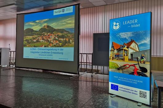 Präsentationsleinwand mit einer Landschaftsaufnahme und dem Text 'ILONA - Onlineveranstaltung in der integrierten ländlichen Entwicklung'. Rechts ein Banner mit dem Logo der LEADER-Initiative und Bildern von Gebäuden.