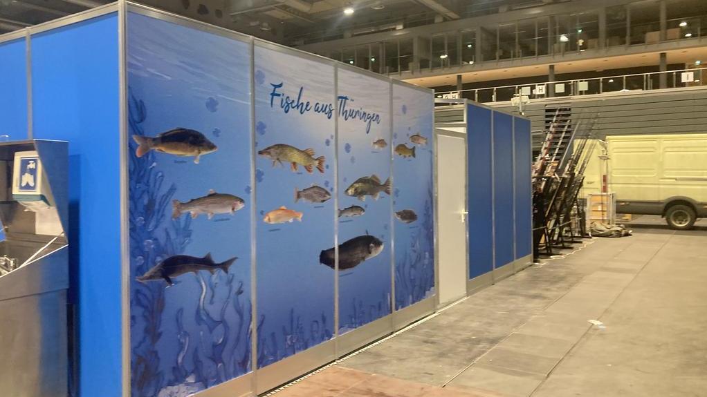 Messestand mit blauen Trennwänden und großflächiger Grafik von Fischen unter Wasser mit der Aufschrift 'Fische aus Thüringen' in einer Halle mit hoher Decke und Industriebeleuchtung.