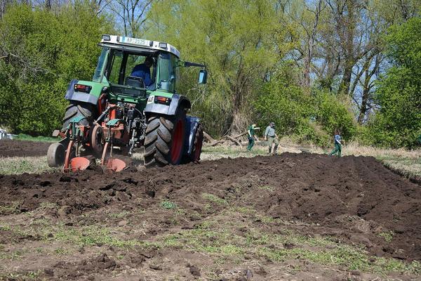 Traktor pflügt ein Feld, im Hintergrund drei Personen mit Arbeitskleidung und Werkzeugen, Bäume am Rand des Feldes.