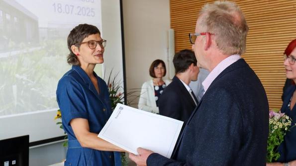 Frau in blauem Kleid erhält von älterem Mann mit Brille und dunklem Sakko ein weißes Dokument in einem Raum mit Holzvertäfelung.