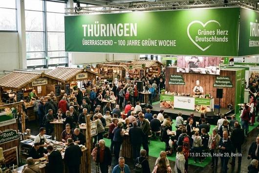 Große Messehalle mit vielen Besuchern an Ständen und Tischen, darüber ein grünes Banner mit der Aufschrift 'THÜRINGEN ÜBERRASCHEND – 100 JAHRE GRÜNE WOCHE' und einem Herzsymbol mit 'Grünes Herz Deutschlands'.