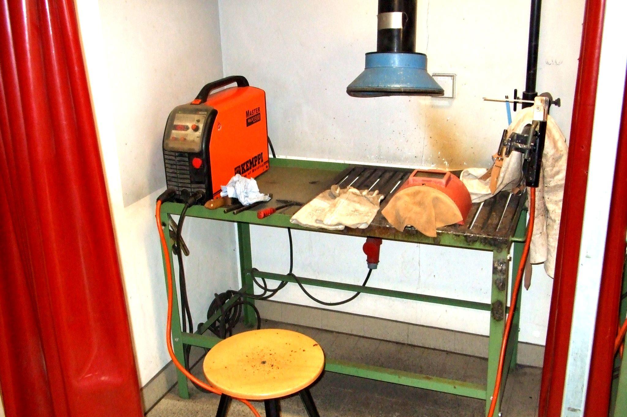 Werkbank mit orangener Schweißgerät, Holzscheibe, Handschuhen und rundem Holzschemel in kleinem Raum mit rotem Vorhang.