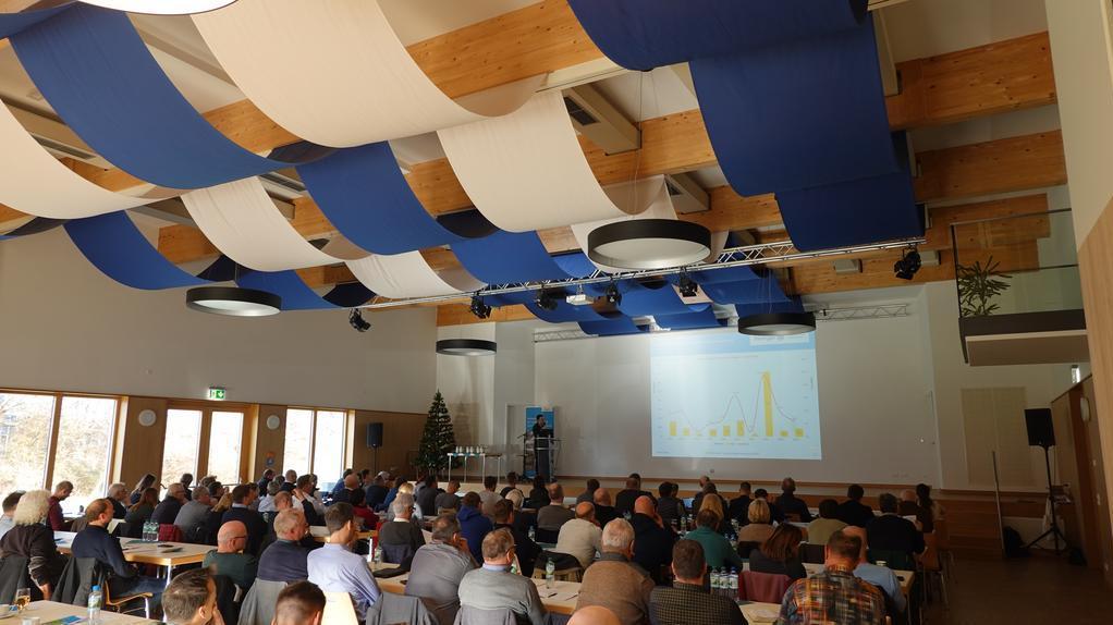 Großer Konferenzraum mit vielen sitzenden Personen, die einer Präsentation mit Diagramm auf einer Leinwand folgen. Decke mit blauen und weißen Stoffbahnen dekoriert.