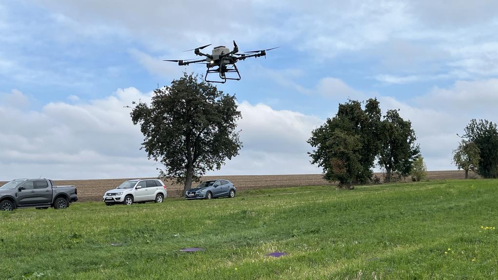 Eine Drohne fliegt über einer grünen Wiese, im Hintergrund stehen drei Autos und mehrere Bäume unter bewölktem Himmel.