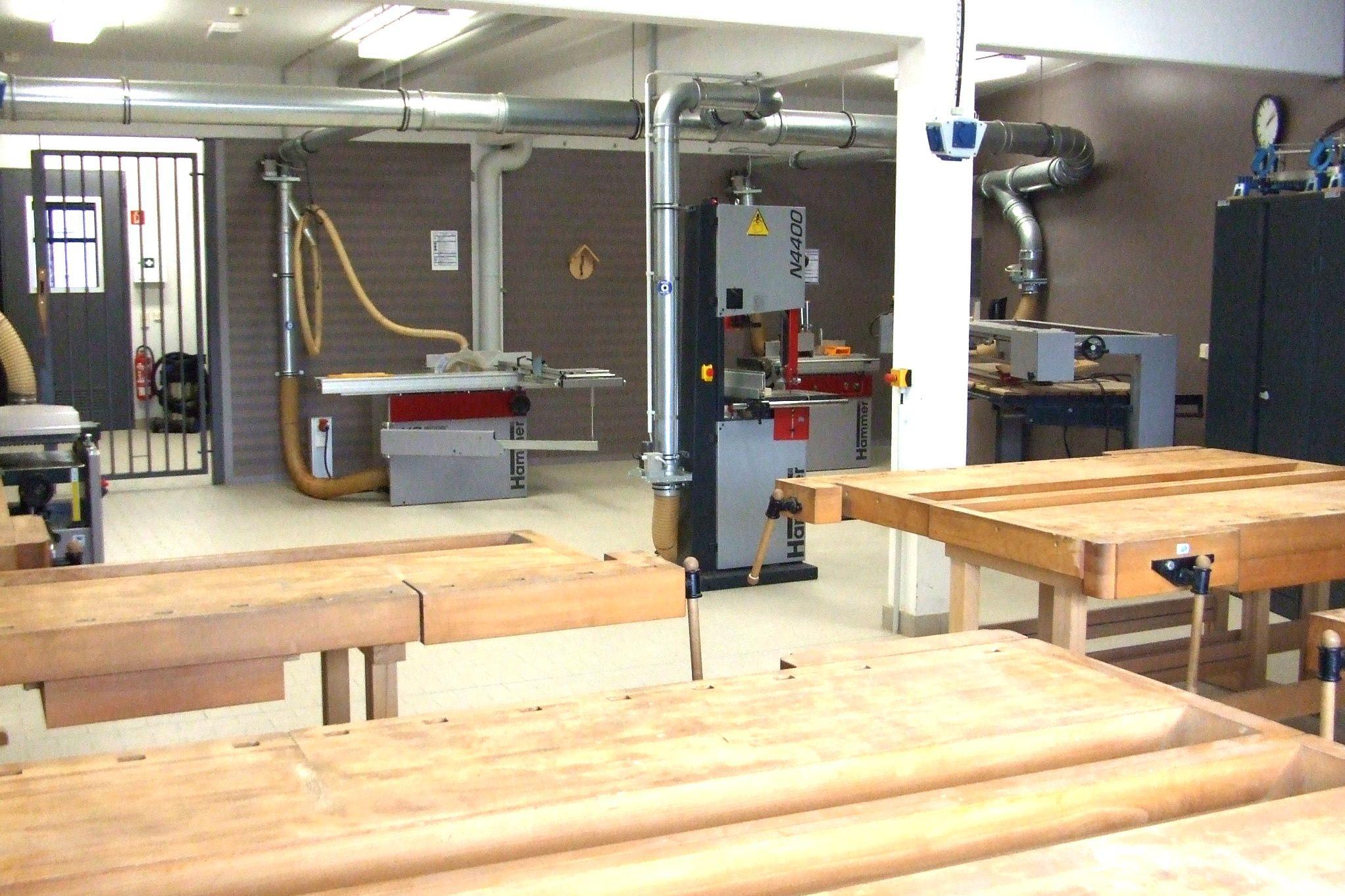 Werkstatt mit Holzbänken, Sägemaschine und Lüftungsrohren an der Decke.