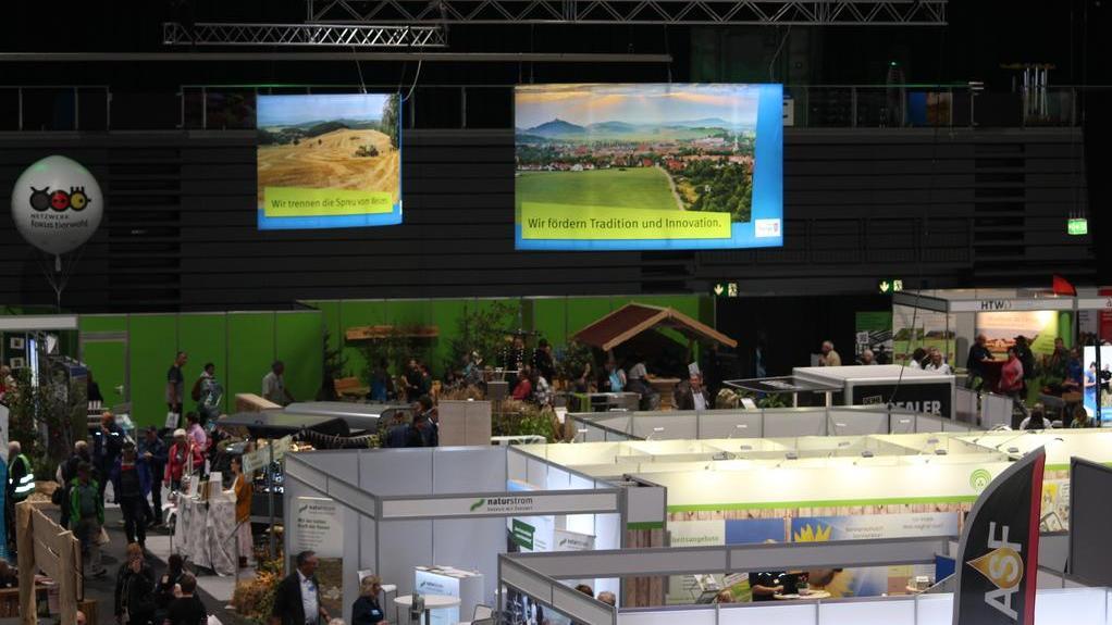 Messestand mit mehreren Ausstellungsflächen und Besuchern in einer großen Halle, darüber zwei große Bildschirme mit Landschaftsfotos und Texten.