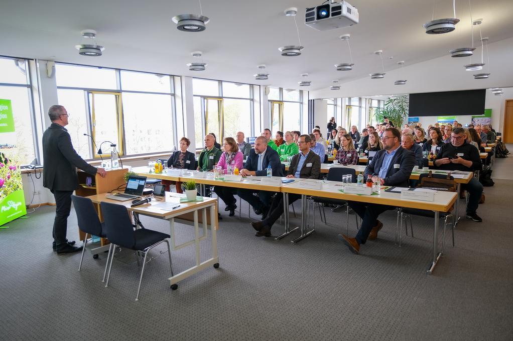 Mann steht an Rednerpult vor Publikum in hellem Konferenzraum mit großen Fenstern und Deckenprojektoren.