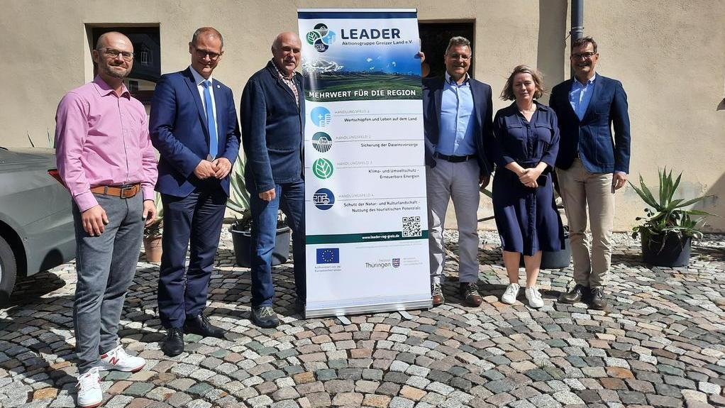 Sechs Personen stehen auf einem gepflasterten Platz vor einem Banner mit dem Text 'LEADER' und weiteren Informationen zur Region. Die Personen tragen formelle Kleidung.