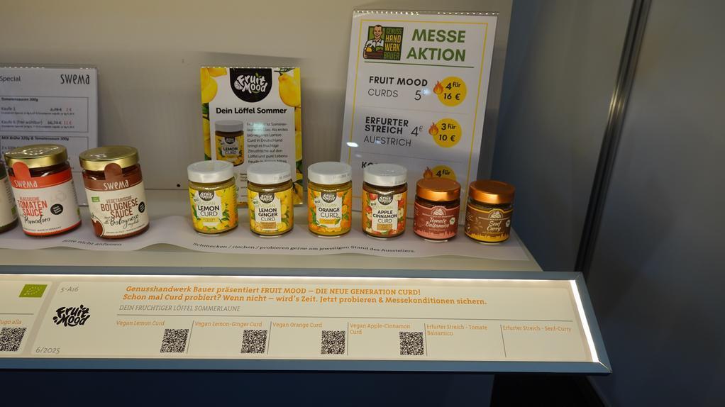 Verschiedene Gläser mit Fruchtaufstrichen und Honig stehen auf einem Messetisch mit Preisschild und Produktbeschreibung.