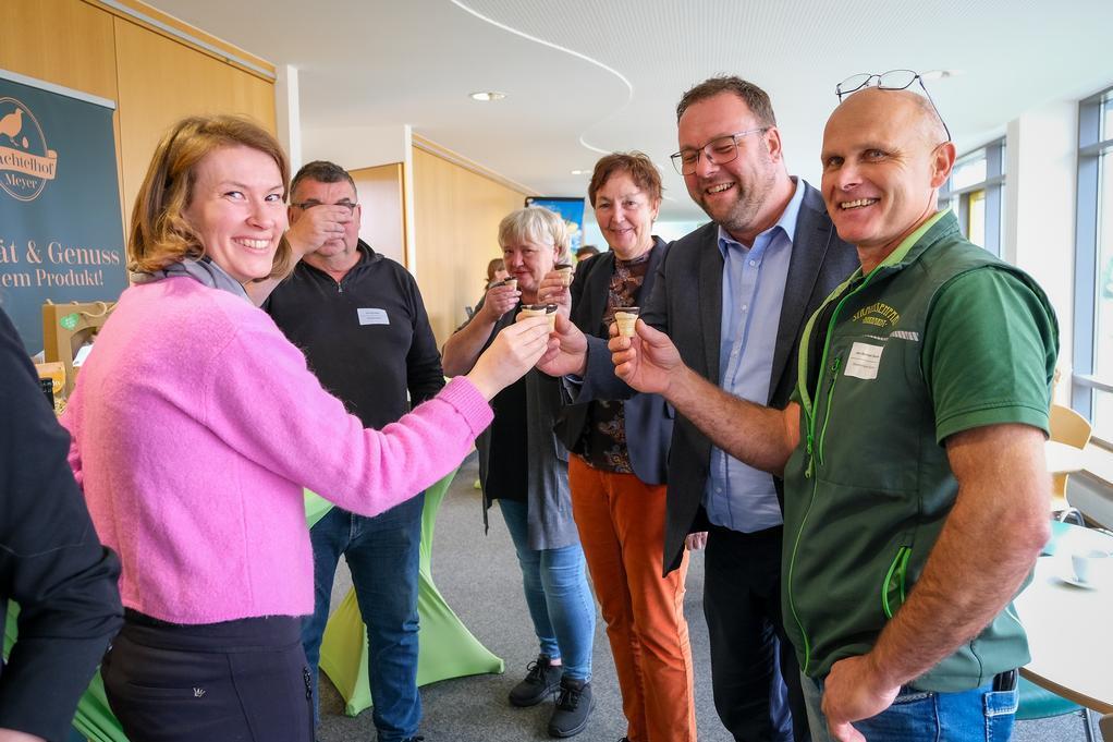 Gruppe von Menschen steht im Kreis und stößt mit kleinen Gläsern an, im Hintergrund Fenster und ein Banner mit Text.