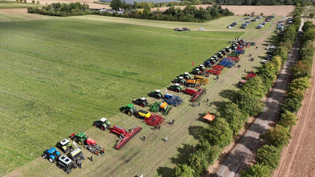 Luftaufnahme von zwei langen Reihen landwirtschaftlicher Fahrzeuge und Geräte auf einem Feld neben einer Baumreihe und einem Feldweg, im Hintergrund Felder und eine Stadt unter bewölktem Himmel.