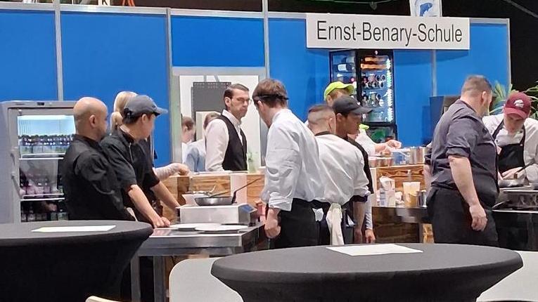 Messestand mit grünem Stehtisch, Informationsdisplays und Personen im Hintergrund in einer Halle