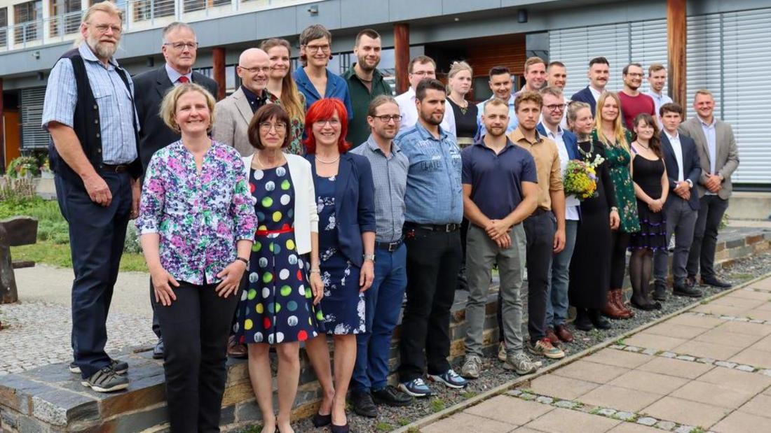 Gruppe von Menschen unterschiedlichen Alters und Geschlechts steht vor einem modernen Gebäude mit großen Fenstern und Balkonen.